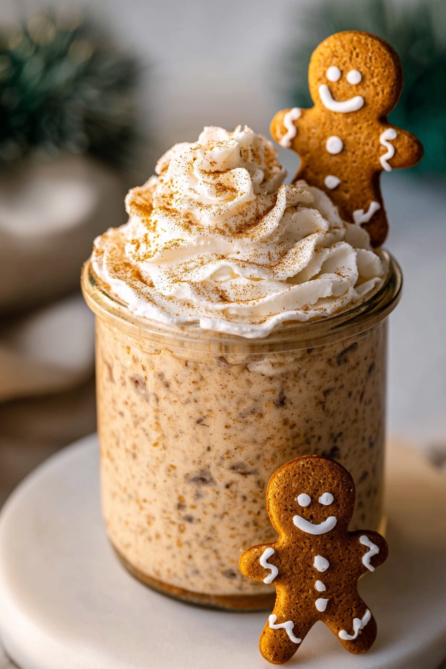 A clear glass jar filled with a thick, tan, speckled mixture that looks creamy and textured, topped with a swirl of white whipped cream sprinkled lightly with cinnamon or nutmeg. A small gingerbread cookie shaped like a smiling person with white icing decoration is placed standing at the rim inside the jar. The jar sits on a white plate on a white marbled surface, with another small gingerbread cookie lying on the plate near the bottom left corner. Blurred green miniature pine trees are in the background. photo taken with an iphone --ar 2:3 --v 7