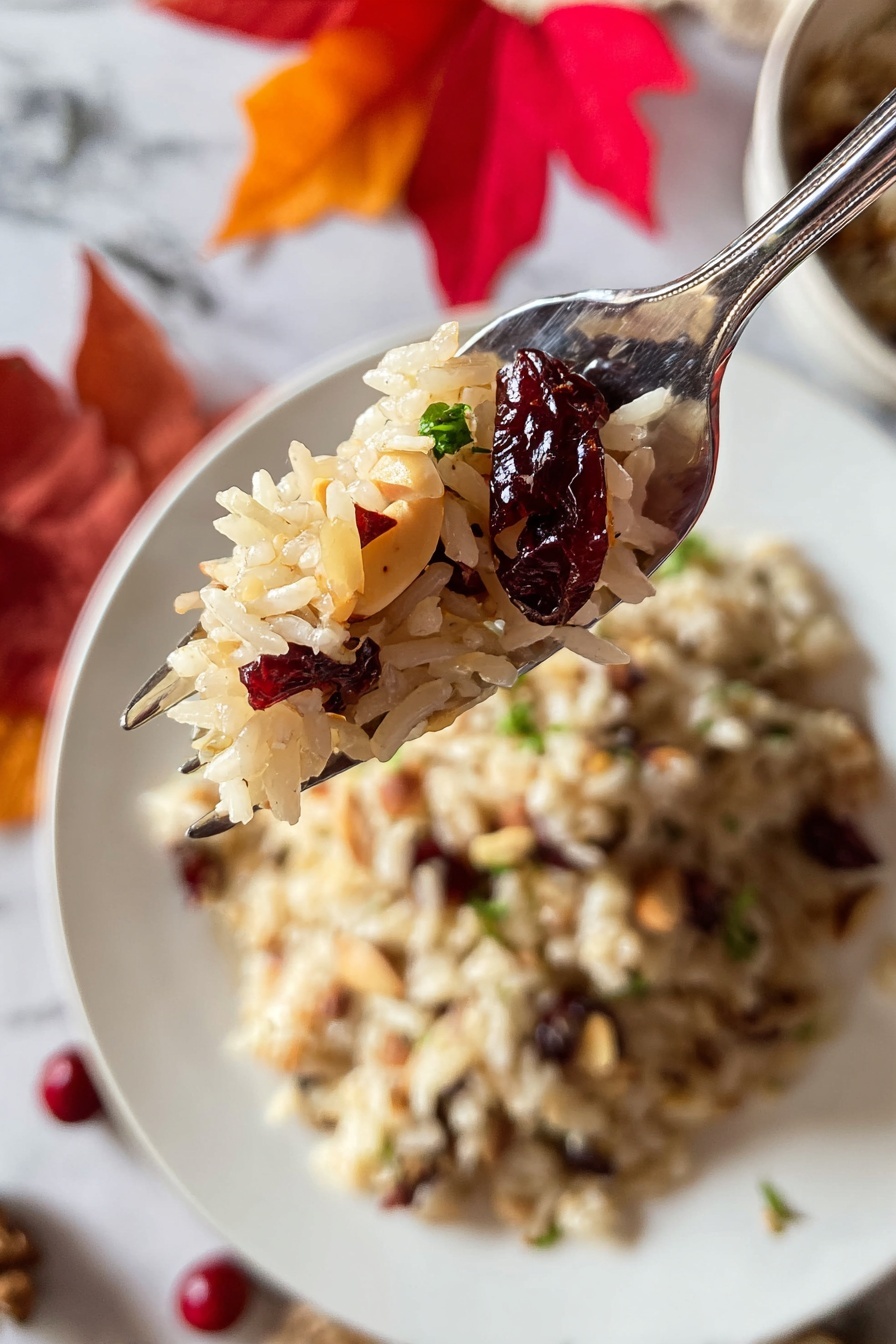 A close-up image shows a silver fork holding a small bite of light brown rice mixed with dry cranberries and light golden slivered nuts. Behind the fork, a white round plate holds a larger serving of the same rice mixture, sprinkled with small green herb pieces. The background features a white marbled surface with scattered red autumn leaves and some herbs around the plate. photo taken with an iphone --ar 2:3 --v 7