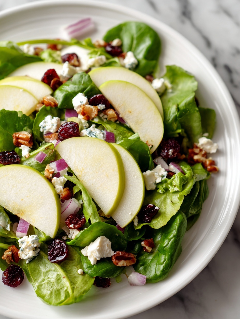 The image shows a fresh salad on a white plate placed on a white marbled surface. The salad has four layers: the bottom layer is a mix of dark green spinach and red-tinted lettuce leaves with a fresh crispy texture; the second layer consists of many thin slices of shiny, bright green apple scattered evenly; the third layer shows small pieces of crunchy walnuts and dried red cranberries spread across the salad; the top layer is dotted with small white crumbles of feta cheese and finely chopped purple onion bits. A silver fork and spoon rest on the left side of the plate. Photo taken with an iphone --ar 2:3 --v 7