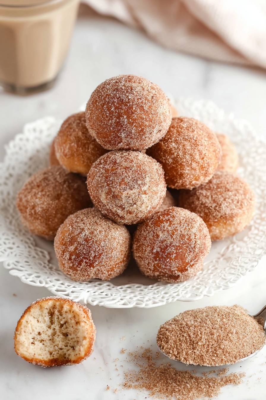 A white plate with a decorative lace-like edge holds a pile of small, round doughnut holes covered evenly in a light brown sugar and cinnamon coating, giving them a grainy texture. One doughnut hole is placed on the white marbled surface in front of the plate, shown whole, while another nearby is broken in half, revealing a soft, fluffy, light beige interior. To the right on the surface, an old silver spoon rests filled with more cinnamon sugar mixture. In the background, partially blurred, there is a glass with a light brown drink and more doughnut holes scattered on the white marbled surface. photo taken with an iphone --ar 2:3 --v 7