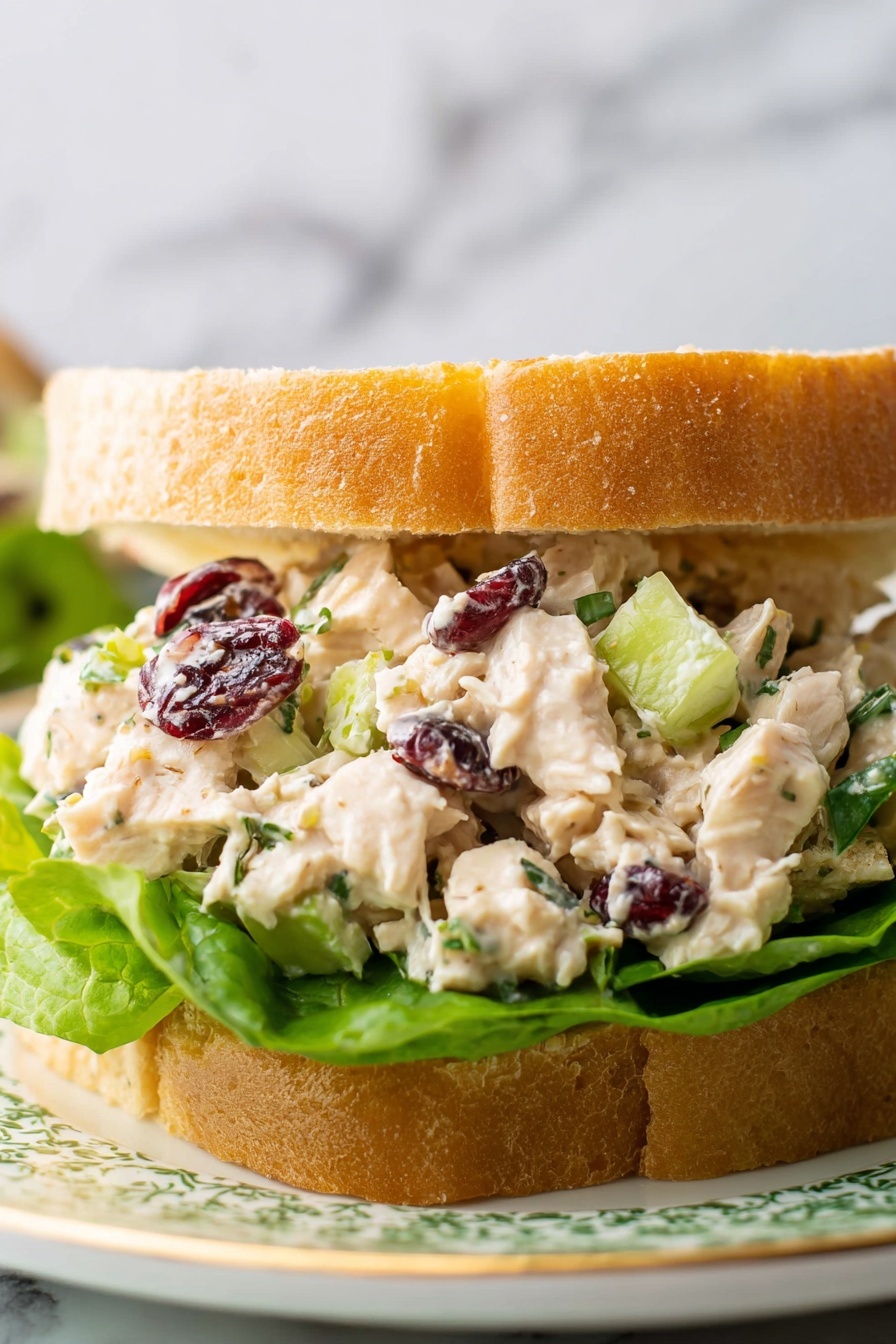 A sandwich with two soft, light brown bread slices forms the base and top layer; inside is a thick, creamy chicken salad mixed with small green celery pieces and dark red dried cranberries, giving it a chunky texture. Fresh, bright green lettuce leaves sit under the chicken mixture, adding a fresh crunch and color contrast. The sandwich rests on a round, white plate with a green striped pattern, placed on a white marbled surface. In the background, a bowl of the same chicken salad is partly visible, along with a colorful striped cloth. photo taken with an iphone --ar 2:3 --v 7