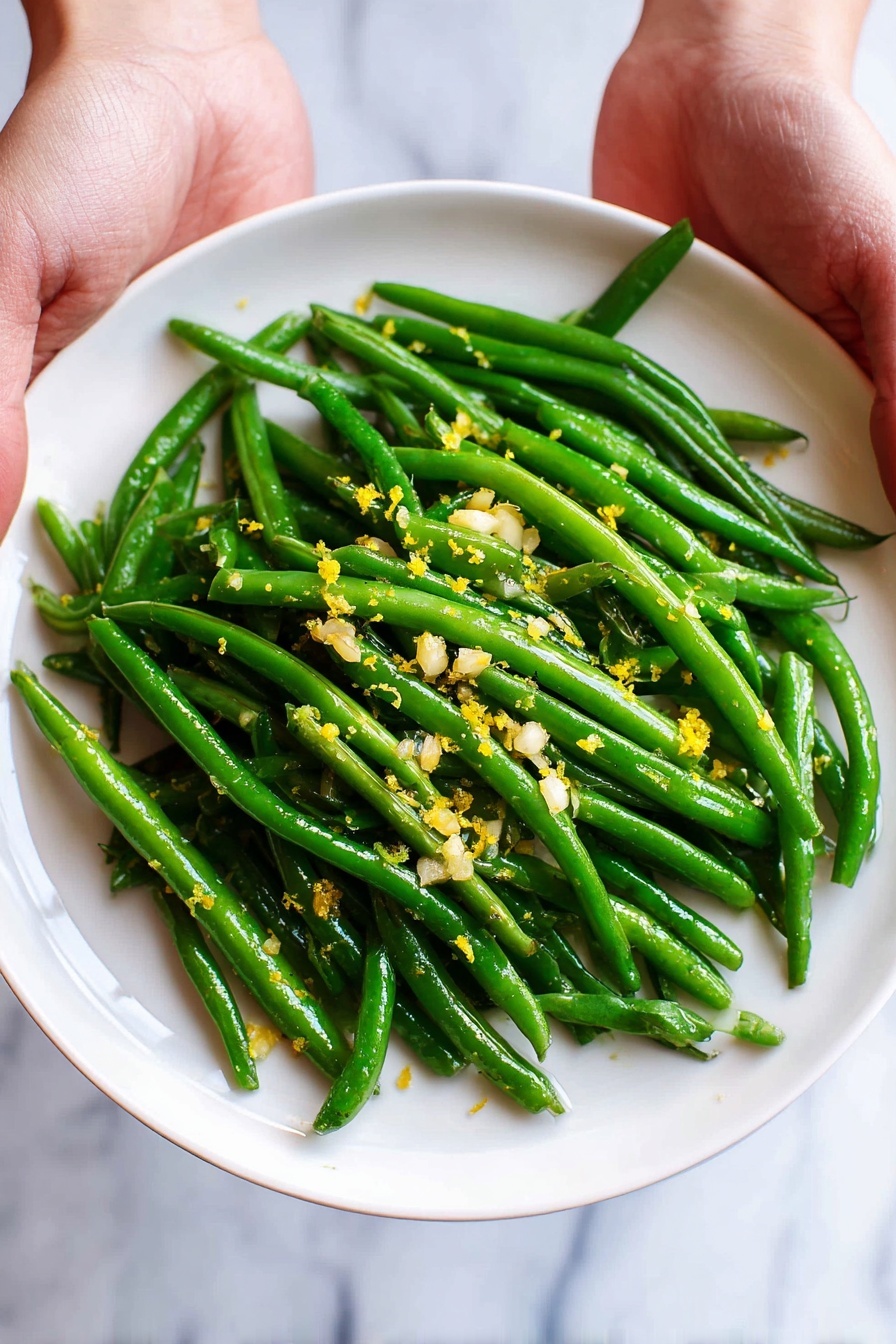 A white plate filled with bright green cooked green beans forms the main layer, arranged in a slightly messy pile. Scattered on top are small pieces of finely chopped garlic and bits of yellow lemon zest, adding texture and color contrast. Two visible woman's hands hold the plate gently from each side, set against a background of white marbled texture. Photo taken with an iphone --ar 2:3 --v 7
