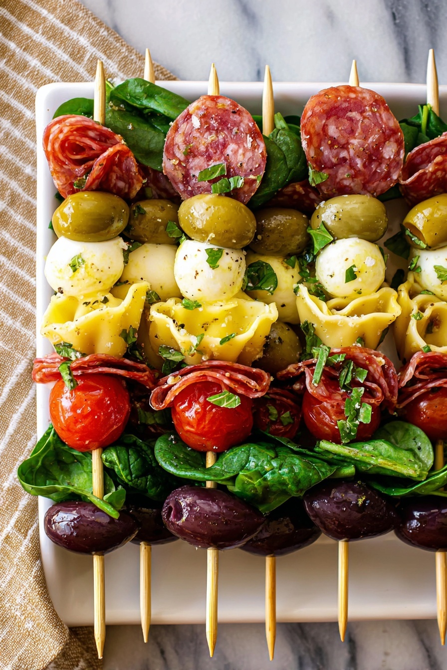 The image shows rows of skewers laid on a black tray, each skewer having five main layers from bottom to top: a large green olive, a folded slice of reddish salami, a small round white mozzarella ball, fresh green spinach leaves, a bright red grape tomato, another folded slice of salami, a yellow cheese tortellini, and a dark purple olive on top. The layers look fresh and glossy with a sprinkle of herbs and seasoning over them. The skewers rest on a green bed of arugula leaves, and the background features a white marbled texture with a beige striped cloth partly visible underneath. Photo taken with an iphone --ar 2:3 --v 7