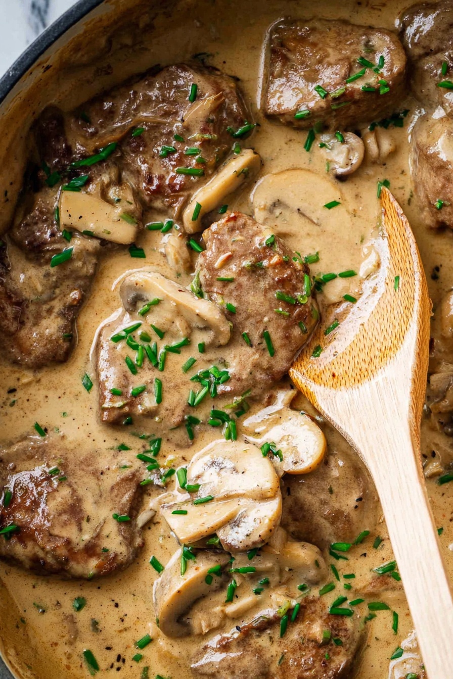 The image shows a creamy dish in a black pan with several brown pieces of cooked meat and light beige slices of mushrooms mixed throughout a thick tan sauce. Small bright green chopped herbs are sprinkled over the top, adding a fresh pop of color. A wooden spoon rests on the right side of the pan, partly covered in the sauce, with a visible smooth, light wood grain texture. The background surface is a white marbled texture. Photo taken with an iphone --ar 2:3 --v 7