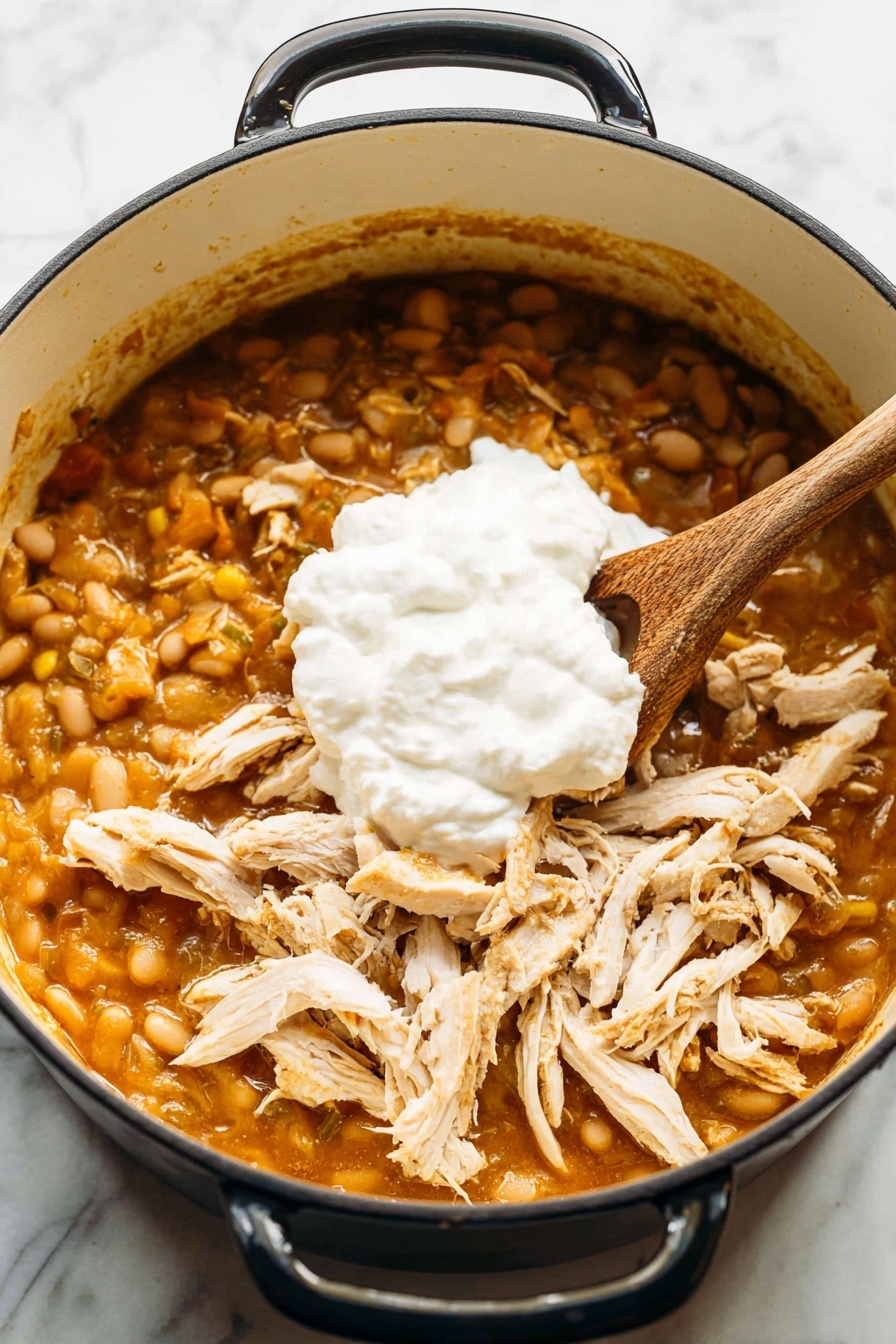 There is a white pot on a white marbled surface filled with a stew that has three visible layers: the bottom layer is a thick yellow bean stew with some orange tint, the middle layer has shredded cooked chicken pieces scattered over the beans, and the top layer is a large dollop of creamy white sauce sitting on the chicken. A wooden spoon is resting inside the pot standing on the left side of the stew. photo taken with an iphone --ar 2:3 --v 7