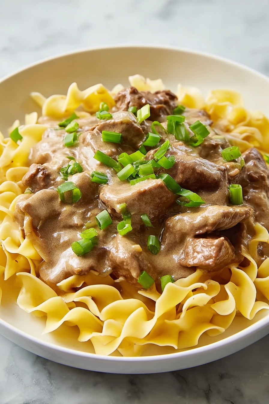 The dish shows curly yellow egg noodles spread as the bottom layer on a white plate, topped with medium-sized strips of beef covered in a thick light brown sauce. On top, scattered sliced green onions add a fresh green contrast, all set against a white marbled surface background. photo taken with an iphone --ar 2:3 --v 7